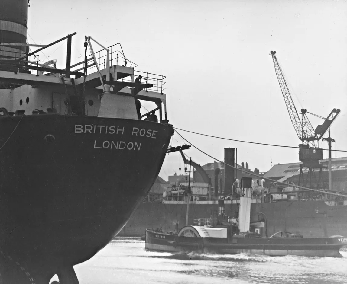 The stern of the tanker British Rose at a fitting out quay on the Wear, with a paddle tug passing and cranes behind.
