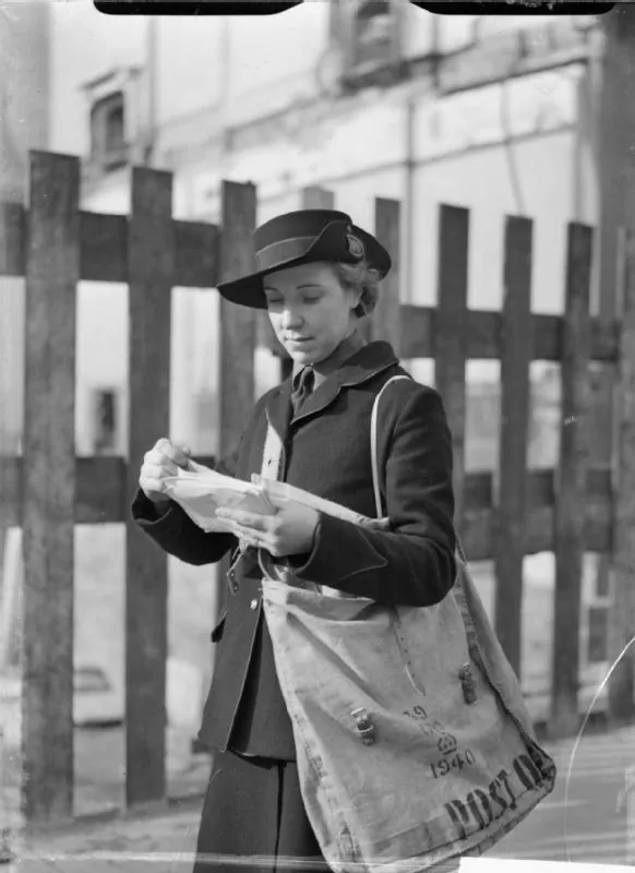 A postwoman with a Post Office bag reads the address on a bundle of letters beside a wooden fence.