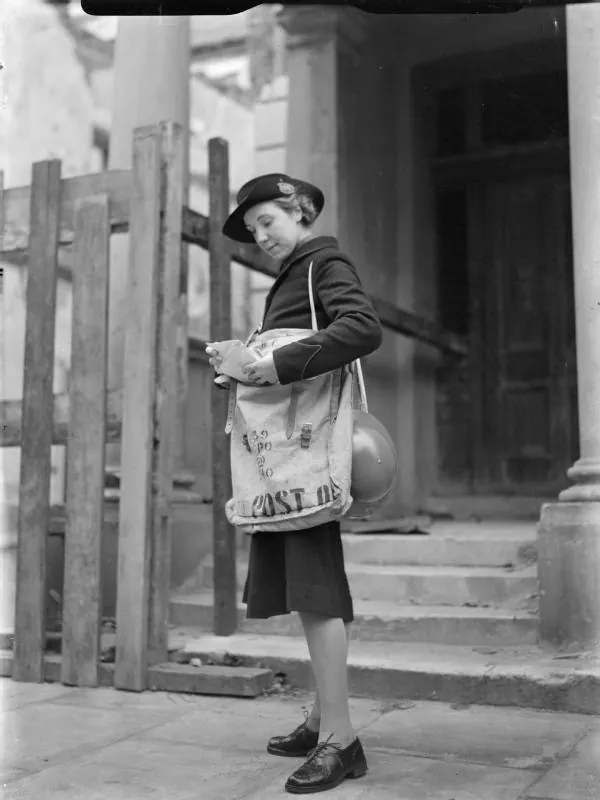 A postwoman stands on a doorstep checking the letters in the Post Office bag slung across her body.