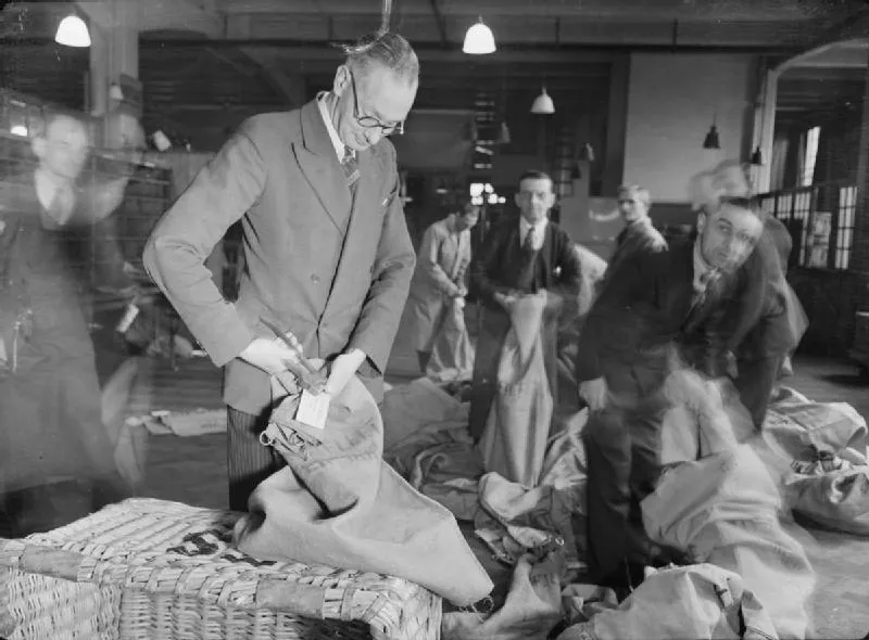A Post Office worker in a suit and glasses seals a mail bag while colleagues fill more bags from wicker baskets behind him.