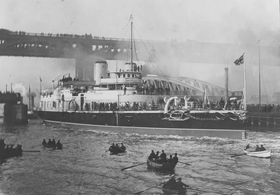 The battleship HMS Victoria steams through the open Swing Bridge at Newcastle, under the High Level Bridge, with rowing boats around her.